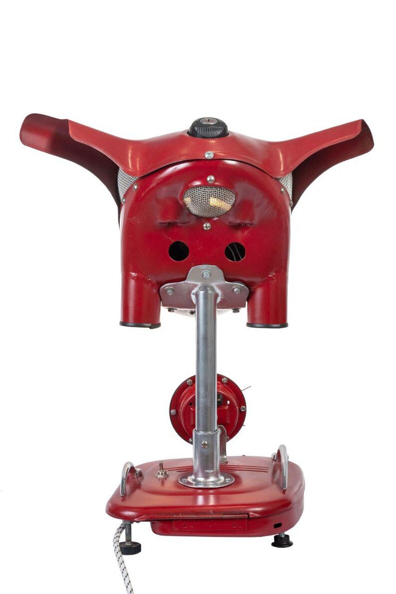 May include: A red vintage electric heater with a unique design resembling a pig's head. The heater has a metal base and a long metal pole that supports the head. The head has two large holes on the sides and a small mesh grill on the front.