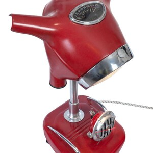 May include: A red table lamp made from a vintage scooter. The lamp has a chrome base and a chrome trim around the top of the lampshade. The lampshade is shaped like a scooter helmet and has a speedometer on the front. The lamp has a chrome horn on the side.