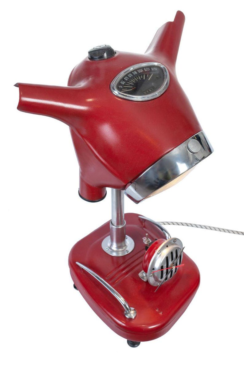 May include: A red table lamp made from a vintage scooter. The lamp has a chrome base and a chrome trim around the top of the lampshade. The lampshade is shaped like a scooter helmet and has a speedometer on the front. The lamp has a chrome horn on the side.