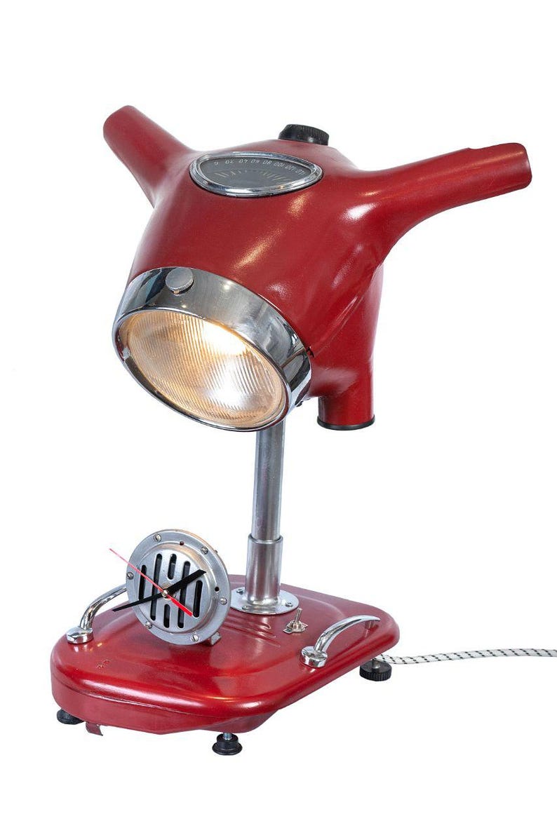 May include: Red and chrome table lamp shaped like a vintage scooter. The lamp has a round headlight, a clock face on the side, and a cord.