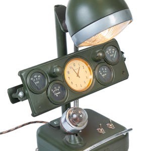 May include: Olive green desk lamp with a vintage aesthetic. Features a domed lampshade, a panel with gauges and a clock, and a base with a small light. The lamp has a retro design with a metallic finish and a decorative power cord.
