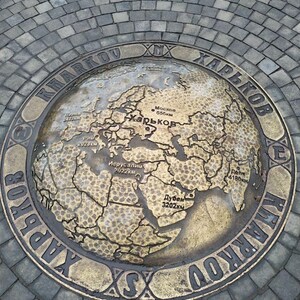 May include: A circular brass plaque with a world map engraved on it. The plaque is surrounded by a ring with the text "EX KHARKOV ХАРЬКОВ" in Cyrillic and Latin script. The map shows distances from Kharkiv to various cities, including Moscow, Rome, Jerusalem, Dubai, and Delhi.