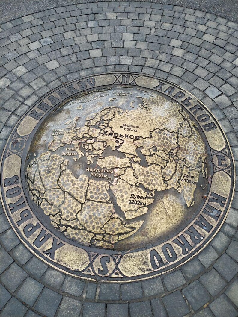 May include: A circular brass plaque with a world map engraved on it. The plaque is surrounded by a ring with the text "EX KHARKOV ХАРЬКОВ" in Cyrillic and Latin script. The map shows distances from Kharkiv to various cities, including Moscow, Rome, Jerusalem, Dubai, and Delhi.