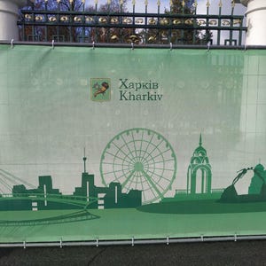 May include: A green banner with the text "Харків Kharkiv" and a silhouette of a city skyline with a Ferris wheel. The banner is hanging on a metal fence.