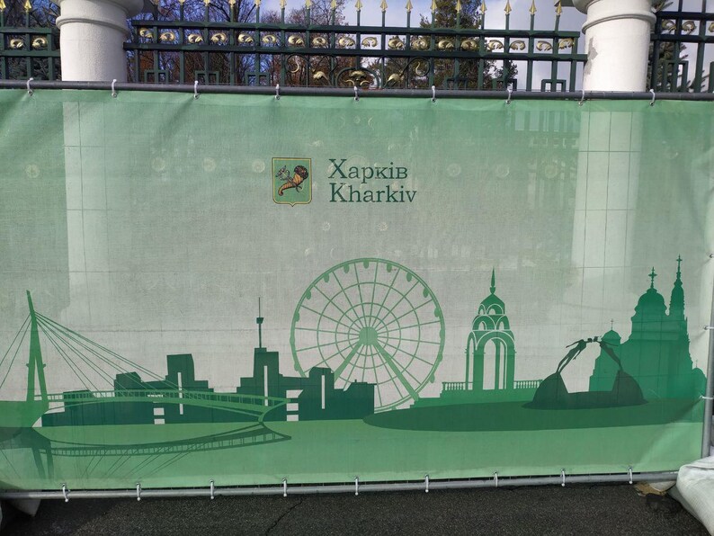 May include: A green banner with the text "Харків Kharkiv" and a silhouette of a city skyline with a Ferris wheel. The banner is hanging on a metal fence.