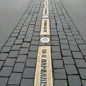 May include: A paved walkway with a series of brass plaques embedded in the ground. The plaques are inscribed with the text "50-я ПАРАЛЛЕЛЬ" in Cyrillic script, which translates to "50th Parallel".
