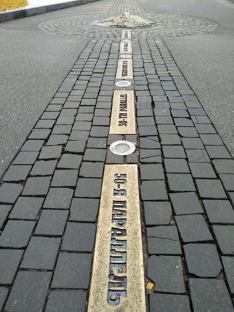 May include: A paved walkway with a series of brass plaques embedded in the ground. The plaques are inscribed with the text "50-я ПАРАЛЛЕЛЬ" in Cyrillic script, which translates to "50th Parallel".