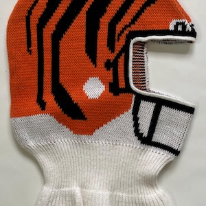 May include: Knitted balaclava in orange and black tiger stripe design, with white accents. The balaclava covers the head and neck, with openings for the eyes and mouth. A winter accessory for cold weather.