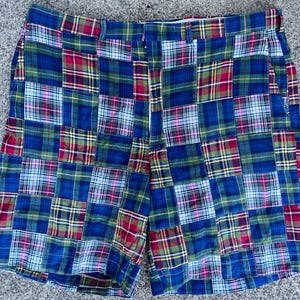 May include: A pair of shorts featuring a patchwork design made of various plaid fabric squares in shades of blue, red, green, yellow, and white. The shorts have a waistband with belt loops, a front fly closure, and side pockets.