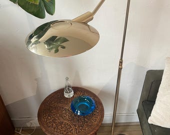 May include: A gold floor lamp with a curved arm and a round, flat shade. The lamp is standing on a white surface next to a small, intricately carved wooden table with a blue glass ashtray.