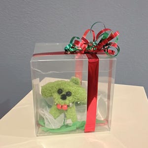 May include: A green plush dog toy inside a clear plastic gift box. The box is decorated with a red ribbon and a red, green, and gold bow. The toy has black eyes, a pink tongue, and a silver collar. The gift box is sitting on a white surface.