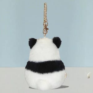 以下が含まれることがあります： 金色のチェーンが付いた、パンダ型のぬいぐるみキーホルダー。パンダは白い体に黒い縞模様と黒い耳があります。キュートで持ち運びやすいアクセサリーです。
