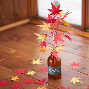 May include: A decorative arrangement featuring paper maple leaves in shades of red, yellow, and orange. The leaves are arranged on a stem inside a brown glass bottle with the text "goel". Scattered leaves are on a wooden surface.