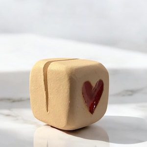 May include: A small, square, beige ceramic card holder with a deep red heart on one side. The holder has a vertical slit on top for inserting cards or photos. The heart is painted with a glossy finish.