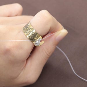 Puede incluir: Un anillo de dedo de color latón con diseños florales y de hojas y la palabra "CLOVER" grabada. El anillo tiene una pequeña cuchilla de color plateado adjunta. Un hilo blanco está siendo sostenido por el anillo.