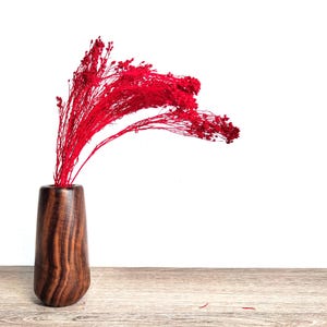 Può includere: Un vaso di legno con finitura marrone scuro contiene una vivace composizione di fiori secchi rossi. Il vaso ha una base arrotondata e una parte superiore leggermente svasata. La composizione è ambientata su uno sfondo bianco e poggia su una superficie di legno chiaro.