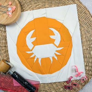 Könnte beinhalten: Ein weißer Stoffquadrat mit einem großen orangenen Kreis in der Mitte. Der Kreis hat eine weiße Krabben-Silhouette im Inneren. Der Stoff liegt auf einer geflochtenen Strohmatte.