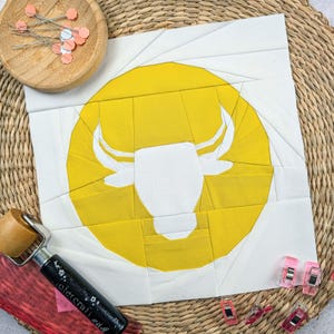 Könnte beinhalten: Eine weiße Bullensilhouette auf einem gelben Kreis, umgeben von weißem Stoff. Der Stoff liegt auf einer geflochtenen braunen Oberfläche.