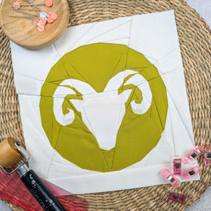 Könnte beinhalten: Ein weißer Stoffquadrat mit einem gelben Kreis in der Mitte. Der Kreis zeigt eine weiße Silhouette eines Widderkopfes mit Hörnern. Der Stoff ist mit rosa und weißen Stecknadeln auf einer gewebten Matte befestigt.