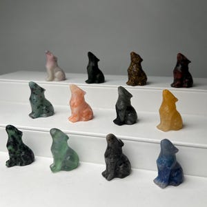 May include: A collection of small, carved wolf figurines in various colors, including pink, black, green, orange, brown, yellow, and blue. The wolves are in a howling pose, and are displayed on a white surface.