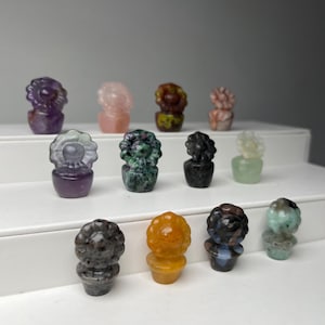 May include: A collection of small, carved gemstone cactus figurines in various colors. The cacti have detailed flower-like tops and are set in small pot-like bases. Colors include purple, pink, green, yellow, and black. The figurines are displayed on a white tiered surface.