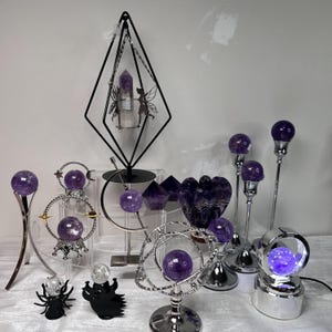 May include: A collection of amethyst crystal spheres and decorative stands. The display includes a black geometric stand with a crystal, a heart-shaped amethyst, and various silver-toned stands with crystal spheres. The spheres vary in size and are a deep purple color.