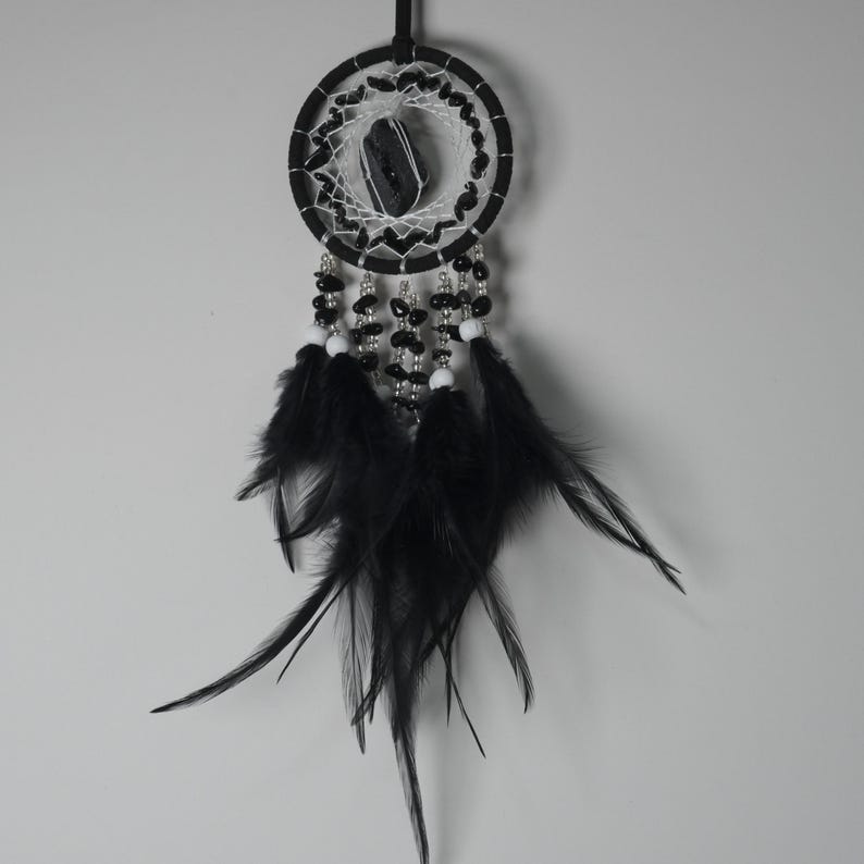 May include: A black and white dreamcatcher with a central stone and hanging feathers. The circular frame is black, with a white web design and black beads. Black feathers hang from the bottom, creating a decorative and spiritual piece.