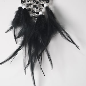 May include: Close-up of a dreamcatcher with black feathers, white beads, and black stone beads. The feathers are long and flowing, and the beads are strung together with silver wire. The dreamcatcher is against a white background.