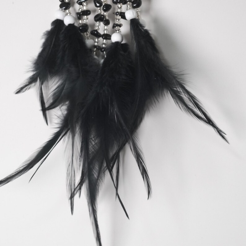 May include: Close-up of a dreamcatcher with black feathers, white beads, and black stone beads. The feathers are long and flowing, and the beads are strung together with silver wire. The dreamcatcher is against a white background.