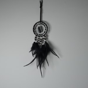 May include: A black and white dreamcatcher with a central stone, beaded accents, and hanging feathers. The dreamcatcher is suspended by a dark brown cord against a neutral gray background. The circular design is a traditional Native American craft.