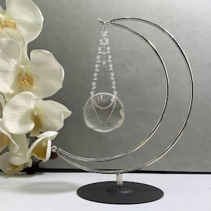 May include: A clear glass orb with silver chains and a beaded strand hangs from a silver metal crescent moon-shaped stand. The stand has a black circular base. White orchids are in the background.