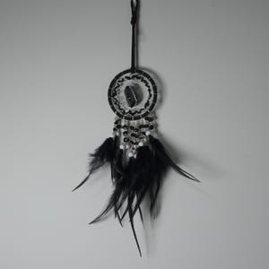May include: A black and white dreamcatcher with a central stone, intricate web design, and hanging black feathers. The dreamcatcher is suspended by a black cord, showcasing a blend of natural and handcrafted elements.