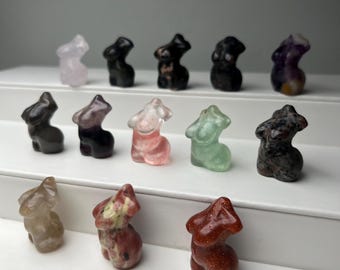 May include: A collection of small, carved gemstone figurines in the shape of a torso. The figurines are in various colors, including pink, green, black, and brown. The collection is displayed on a white shelf.