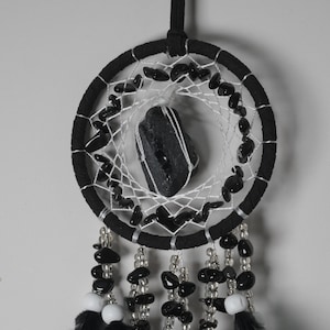 May include: A black dreamcatcher with a central black stone and a web of white string. The circular frame is wrapped in black fabric and adorned with black beads. Strands of beads and feathers hang from the bottom.