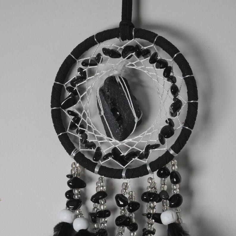 May include: A black dreamcatcher with a central black stone and a web of white string. The circular frame is wrapped in black fabric and adorned with black beads. Strands of beads and feathers hang from the bottom.