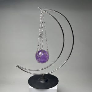 May include: A decorative amethyst sphere suspended by a silver chain within a crescent-shaped metal stand. The sphere is a vibrant purple color, and the stand has a black base. The design is elegant and minimalist.