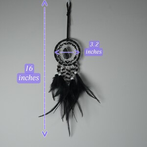 May include: A black dreamcatcher, 16 inches long and 3.2 inches wide, hangs against a gray background. The dreamcatcher features black feathers, beads, and a woven center. The design includes a black cord for hanging.