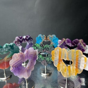 May include: A collection of heart-shaped gemstone sculptures in various colors, including purple, green, blue, and yellow. Each heart is mounted on a small silver stand. The gemstones have unique patterns and textures.