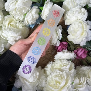 Könnte beinhalten: Weißer Selenit-Stab mit sieben Chakra-Symbolen, die in Regenbogenfarben graviert sind. Die Symbole sind vertikal von unten nach oben angeordnet.