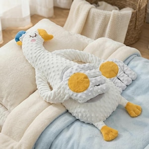 May include: A white goose-shaped cushion with a blue cap and yellow beak and feet. The goose has a textured body and large yellow circles on its wings. It rests on a light blue blanket and cream-coloured pillows.