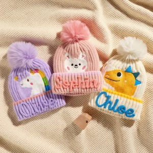 Pode incluir: Três gorros de inverno de malha com pompons. Um é lavanda com um unicórnio e arco-íris, outro é rosa com um coelho e o terceiro é creme com um dinossauro. Cada chapéu tem um nome bordado na aba.