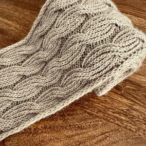 Puede incluir: Un gorro de punto gris claro con un diseño de punto trenzado. El gorro está hecho de un hilo suave y texturizado y está doblado en el borde. Está sobre una superficie de madera.