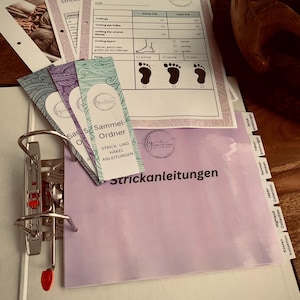 Puede incluir: Una carpeta con una cubierta morada y el texto "Strickanleitungen" impreso en ella. La carpeta tiene un mecanismo de anillas metálicas y está llena de marcadores y papeles de colores. Los papeles están en alemán e incluyen una tabla para medir los pies para tejer calcetines.