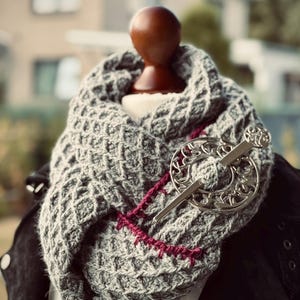 May include: A gray knitted infinity scarf with a textured pattern, accented with a burgundy stitch detail and a silver Celtic-style brooch. The scarf is displayed on a mannequin, showcasing its design and texture.