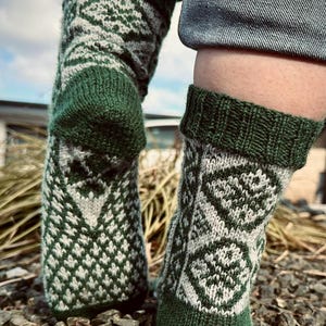 Könnte beinhalten: Ein Paar grüne und weiße Stricksocken mit geometrischem Muster. Die Socken werden getragen und liegen auf einem Bett aus kleinen Steinen.