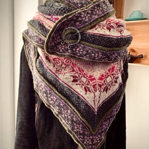 May include: A triangular knitted scarf with a green, pink, gray, and white pattern. The scarf is folded and draped over a mannequin wearing a black leather jacket. A silver metal pin is holding the scarf in place.