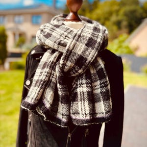 May include: A black and white plaid scarf with a textured knit pattern, draped around a mannequin neck. The scarf features a repeating checkered design with a subtle thin purple line detail along the bottom edge, layered over a black jacket.