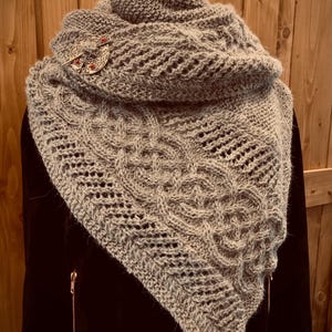 Puede incluir: Un chal gris de punto con un patrón de punto de cable y un broche decorativo. El chal está drapeado sobre un maniquí.