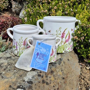 May include: Three white ceramic pots with handles, decorated with floral and bee designs. A small tag reads "Small is Beautiful Sussex Seed Balls". A small cloth bag is next to the pots. The pots are on a rock.
