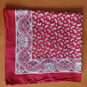 May include: A folded red bandana with a white paisley pattern. The bandana has a detailed border design with floral and scroll elements. The paisley pattern is repeated across the main field of the bandana.